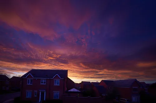 This urban landscape photograph captures a suburban street in Chesterfield during an evening sunset in the winter season. The main subject of the image is a suburban sunset, with a vibrant, colorful sky dominating the scene and displaying hues of orange and purple as the sun sets. The photograph shows rows of brick houses lining the quiet street, emphasizing the intersection of nature and urban environments. The sky creates a dramatic backdrop, highlighting the natural beauty present even within a developed area.